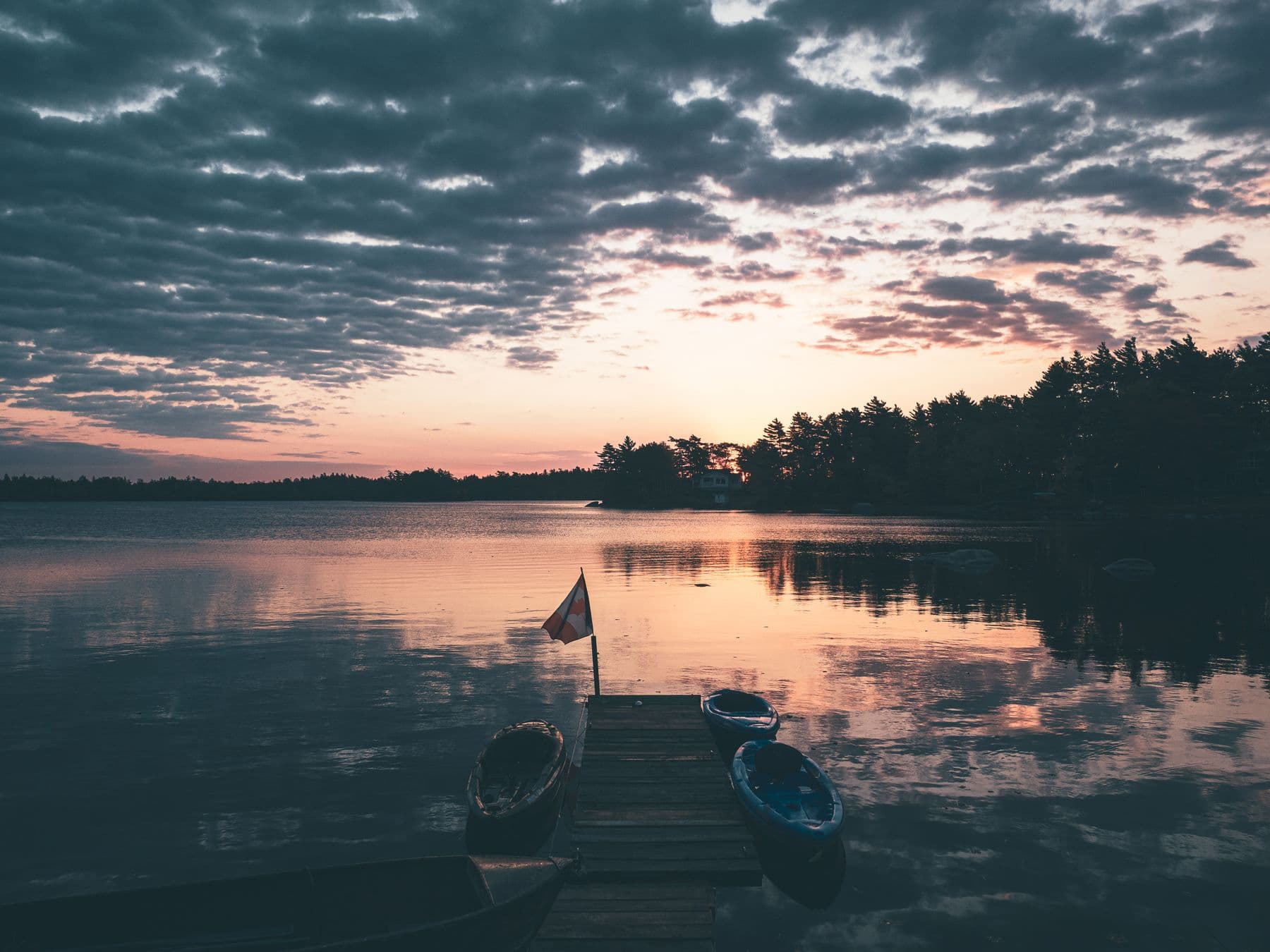 A wooden dock with canoes and a small Canadian flag on a calm lake at sunset
